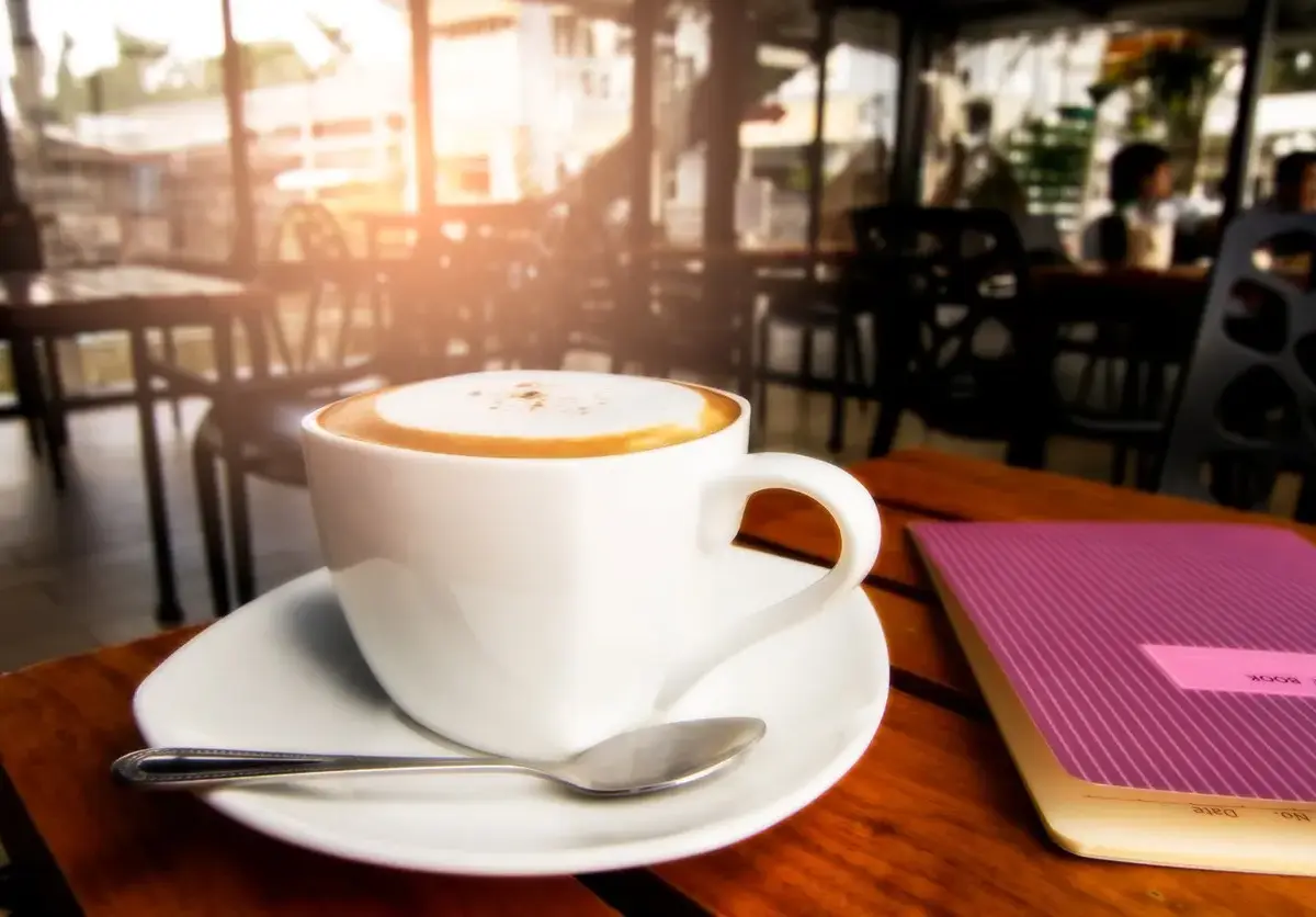 Close-up of a cup of coffee with frothy milk, resting on a saucer next to a notebook in a cozy cafe setting.