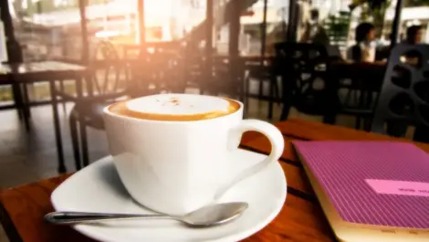 Close-up of a cup of coffee with frothy milk, resting on a saucer next to a notebook in a cozy cafe setting.