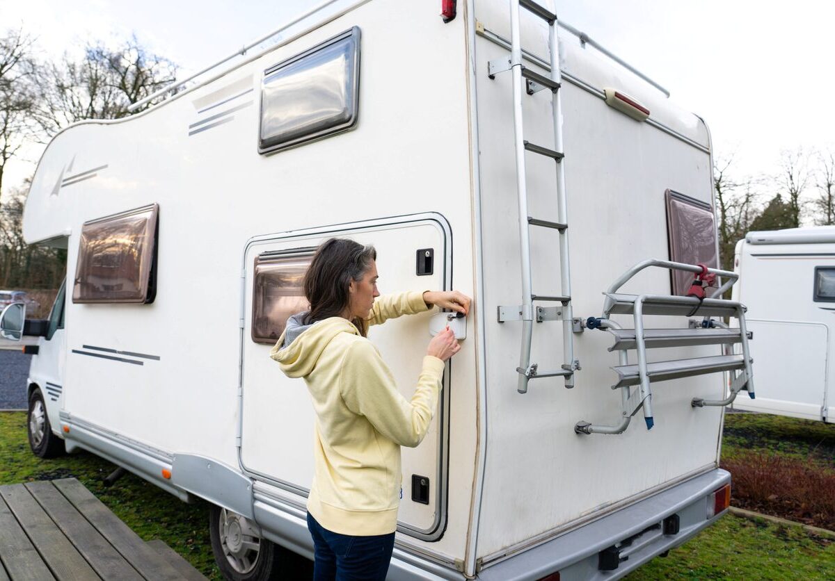 A woman in a yellow hoodie locking the rear door of a white camper van parked at an RV site during the daytime.