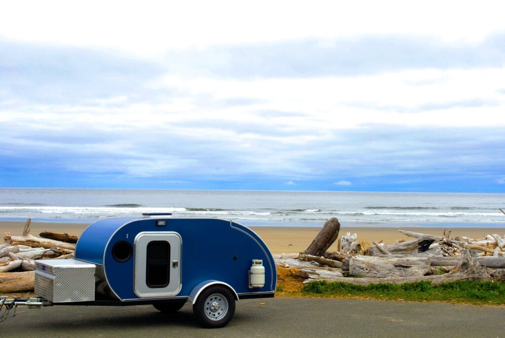 A small blue teardrop trailer parked by a driftwood-covered beach with ocean waves and cloudy skies.
