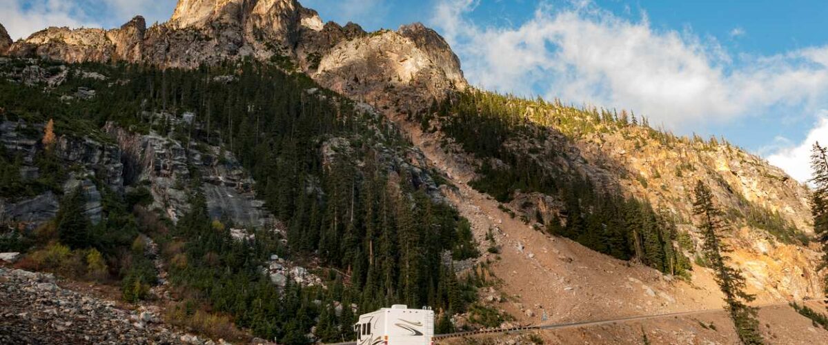An RV traveling through a winding mountain road with rugged cliffs and pine trees under a bright blue sky.