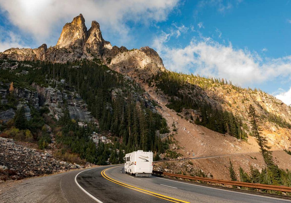An RV traveling through a winding mountain road with rugged cliffs and pine trees under a bright blue sky.