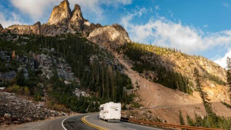 An RV traveling through a winding mountain road with rugged cliffs and pine trees under a bright blue sky.