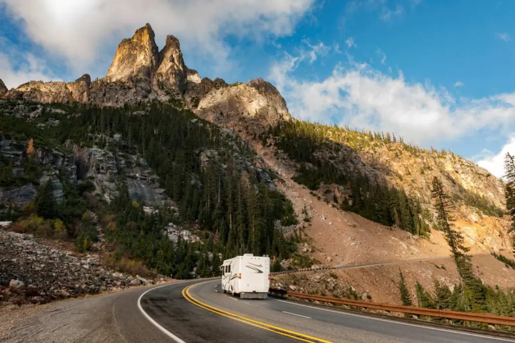 An RV traveling through a winding mountain road with rugged cliffs and pine trees under a bright blue sky.