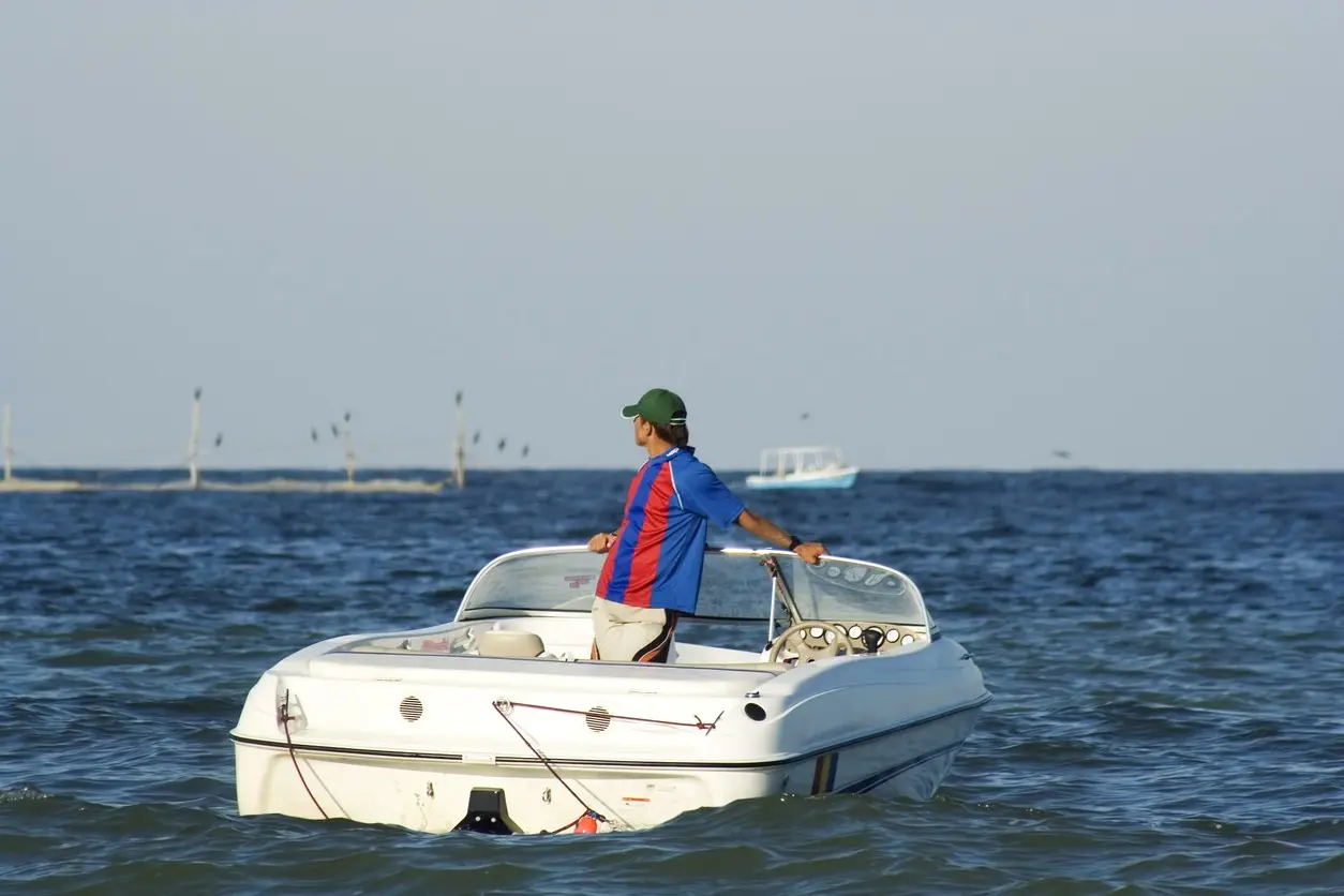 A man in a blue and red shirt standing on a white motorboat, looking out over the open water.