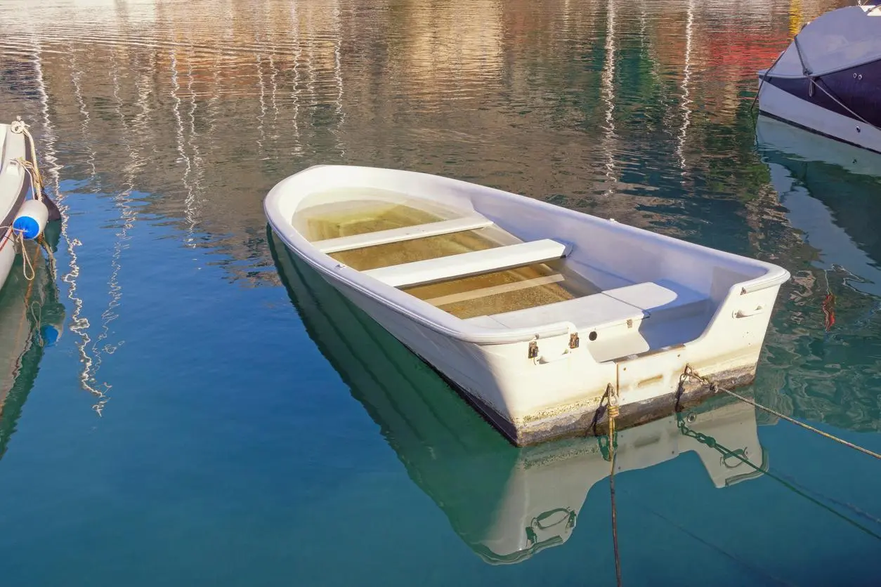 A white rowboat partially filled with water, tied to a dock, and floating on reflective blue water.