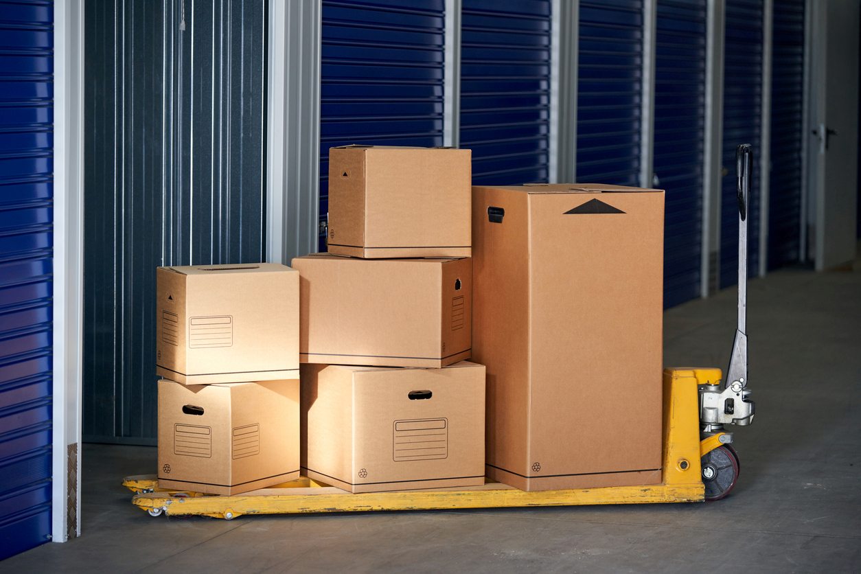 Stacked moving boxes on a yellow pallet jack inside a storage facility with blue roll-up doors.