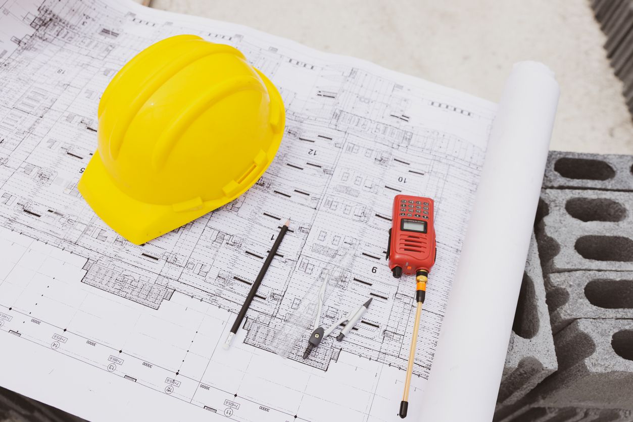 A yellow hard hat, a walkie-talkie, and drafting tools are placed on top of detailed construction blueprints.