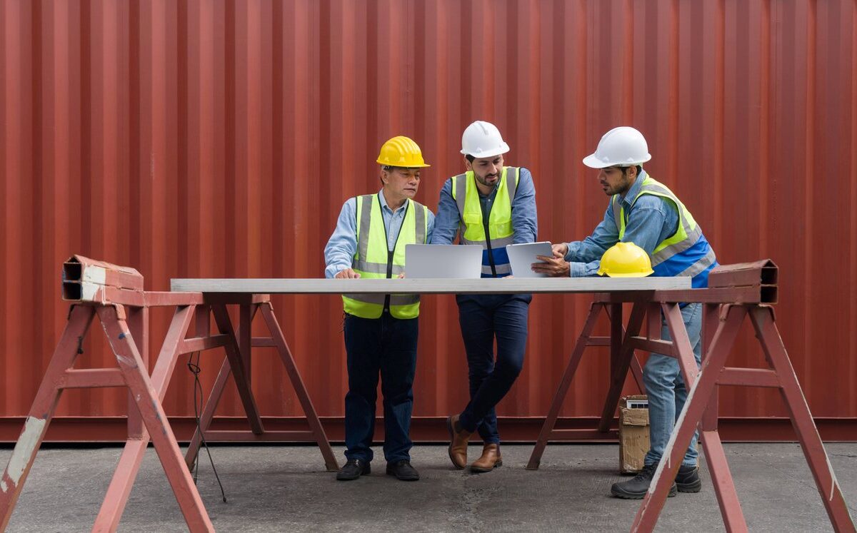 Three construction workers in safety gear review blueprints at a table in front of a large red storage container.