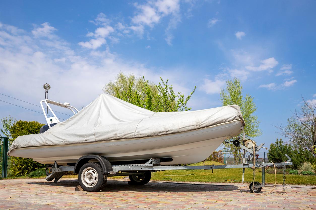 A boat covered with a protective tarp sitting on a trailer in an outdoor storage area under a bright, clear sky.