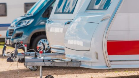 A row of parked RVs and travel trailers lined up outdoors on gravel, showing hitches and front panels.