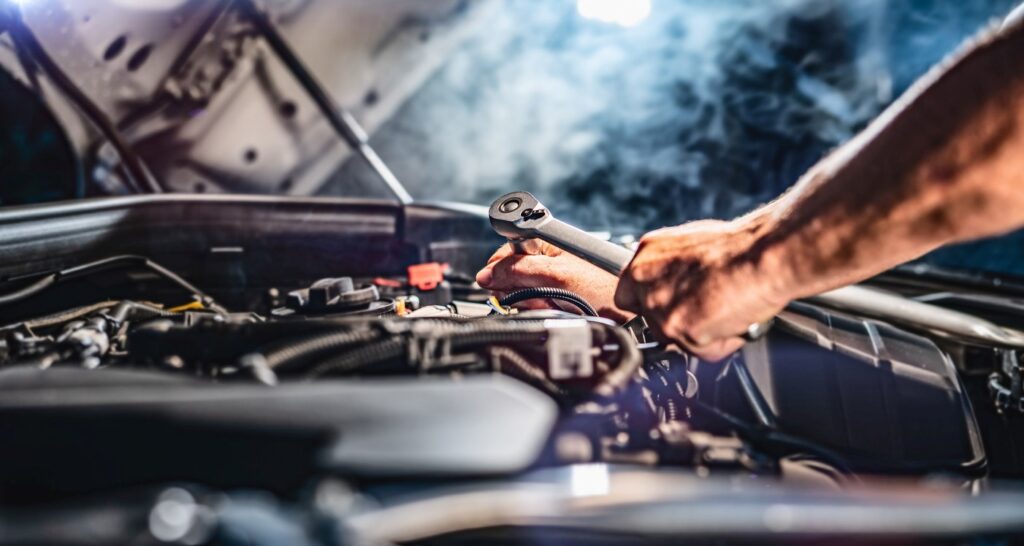 An auto mechanic working on the engine of a car in a garage with steam behind.